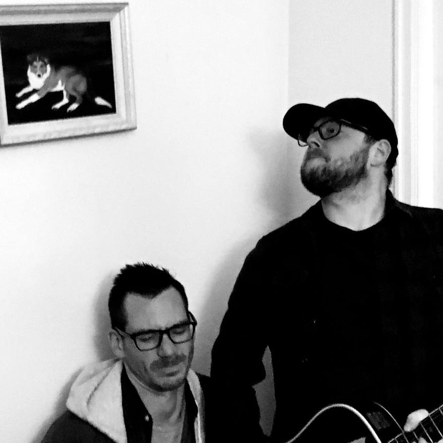 One host playing guitar while the other sits beside him